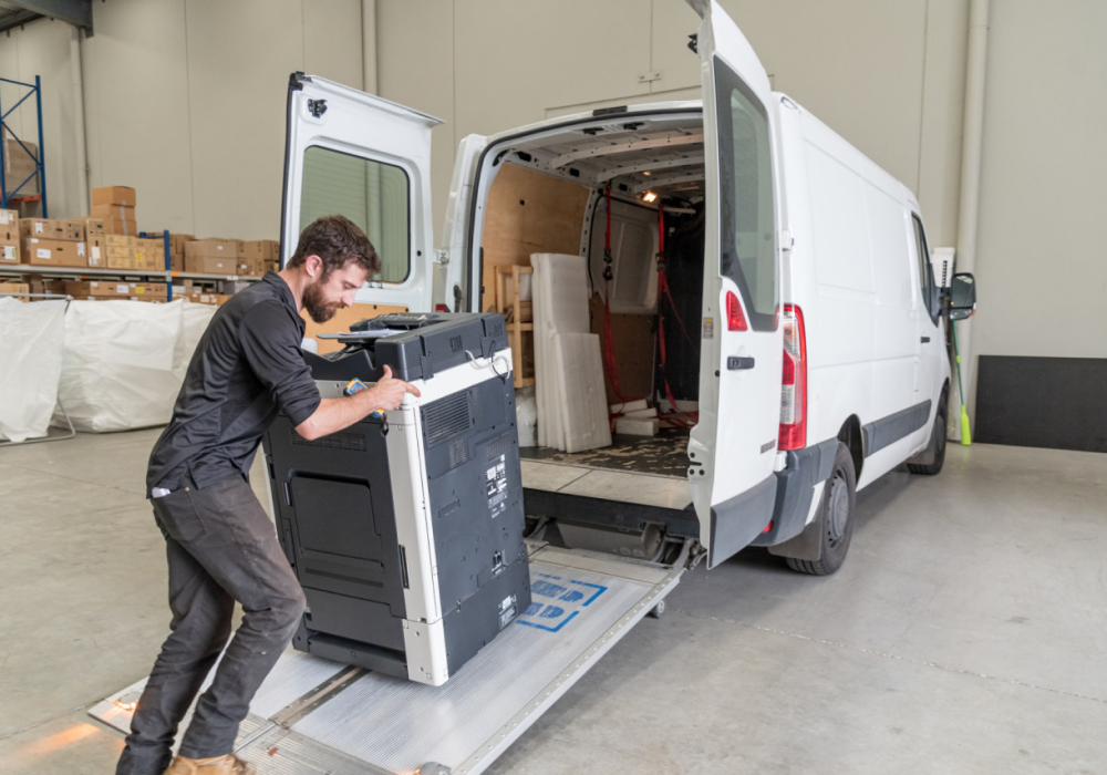 Man loading a large printer into the back of a white van in a warehouse
