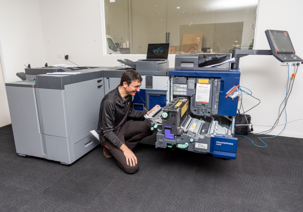 Technician kneeling and servicing a Konica Minolta AccurioPress printer in an office