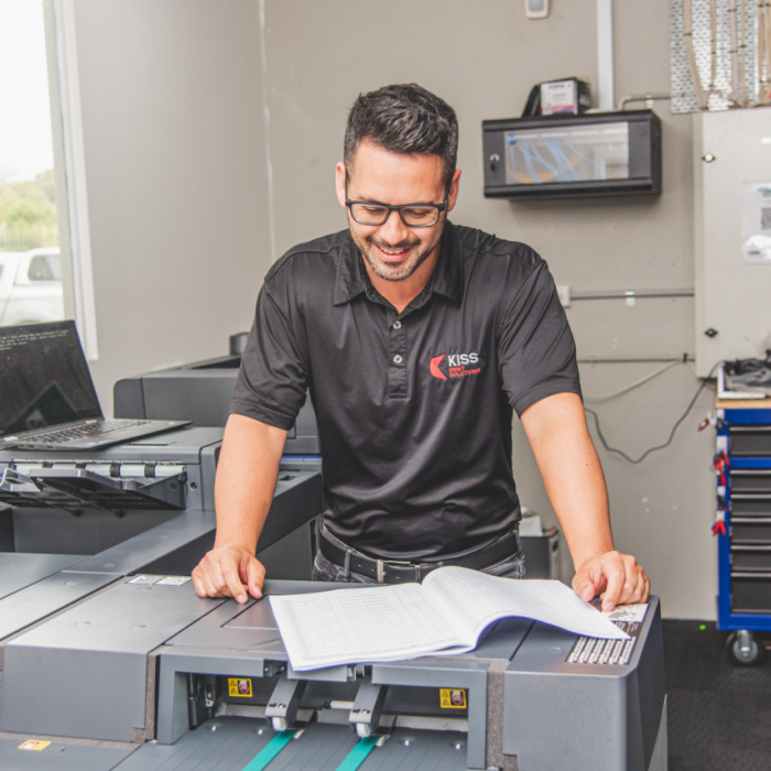 Man in a black KISS Print Solutions shirt reviewing documents at a printer